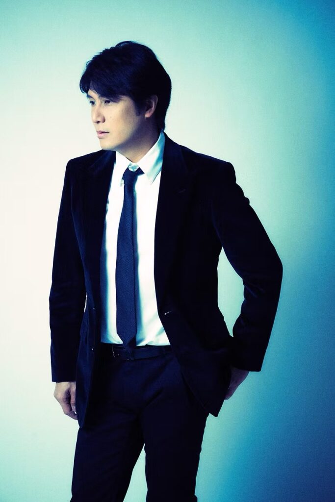 Eisuke Mochizuki in a dark suit against a blue backdrop